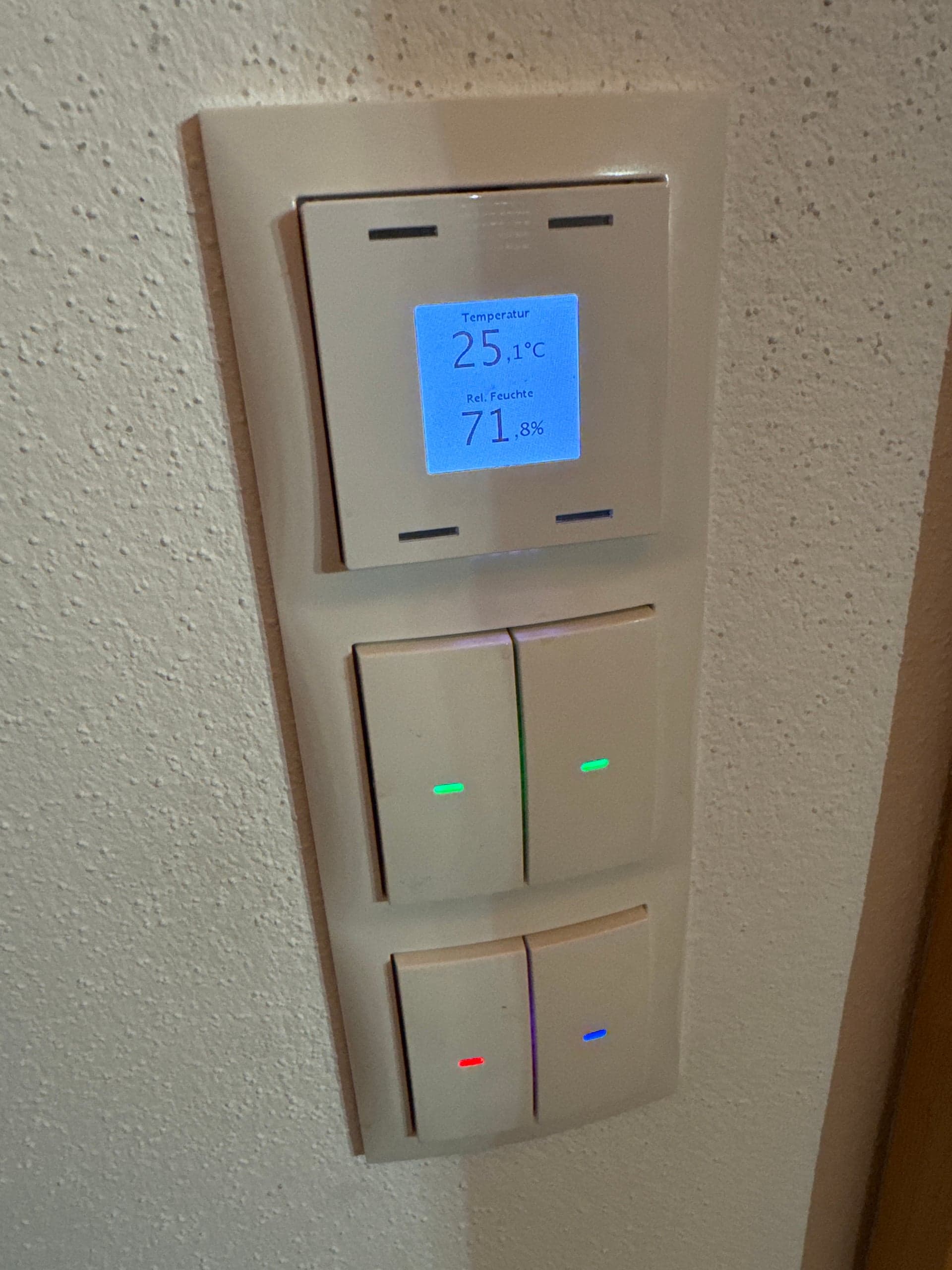KNX-Raumsensor an einer weißen Wand zeigt Temperatur und Luftfeuchtigkeit in einem modernen Wohnraum an